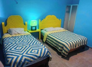Una habitación con dos camas, cada una con una colcha de estampado geométrico. Entre las camas, un mesita de noche con una lámpara verde.