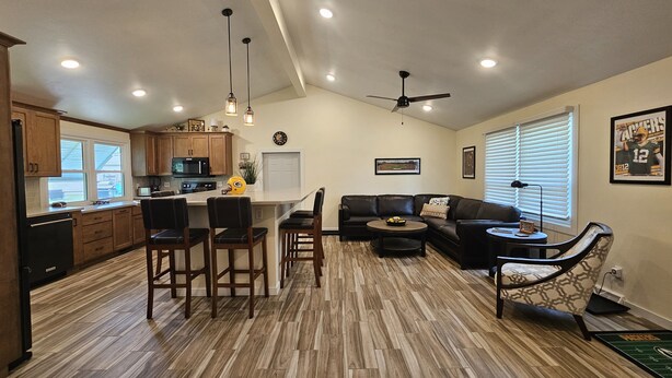 A modern kitchen with wooden cabinets, a central island with bar stools, a dining area with a black leather couch, and a ceiling fan. - Packer themed, renovated 3 bedroom, 2 1/2 bath home- Lambeau Field 16 min walk. (Green Bay)