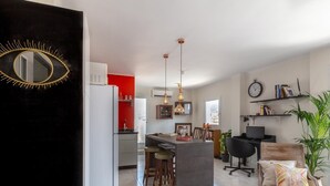 Ett modernt kök med en central köksö, en matplats med en trästol och ett vardagsrum med en TV och hyllor.