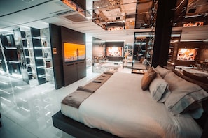 Een moderne hotelkamer met een groot bed, een televisie, een open haard en een glazen scheidingswand met planken.