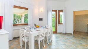 A dining area with a white table set for four, white chairs, a red vase, and a window with a view of greenery. - Rei Sole (REI308) by Interhome (Costa Rei)