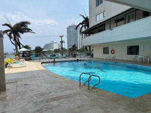Piscina com espreguiçadeiras e um salva-vidas, cercada por palmeiras e edifícios.