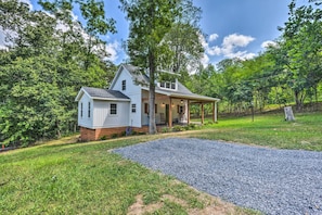 A single-story house with a covered porch, surrounded by lush greenery and a gravel driveway.