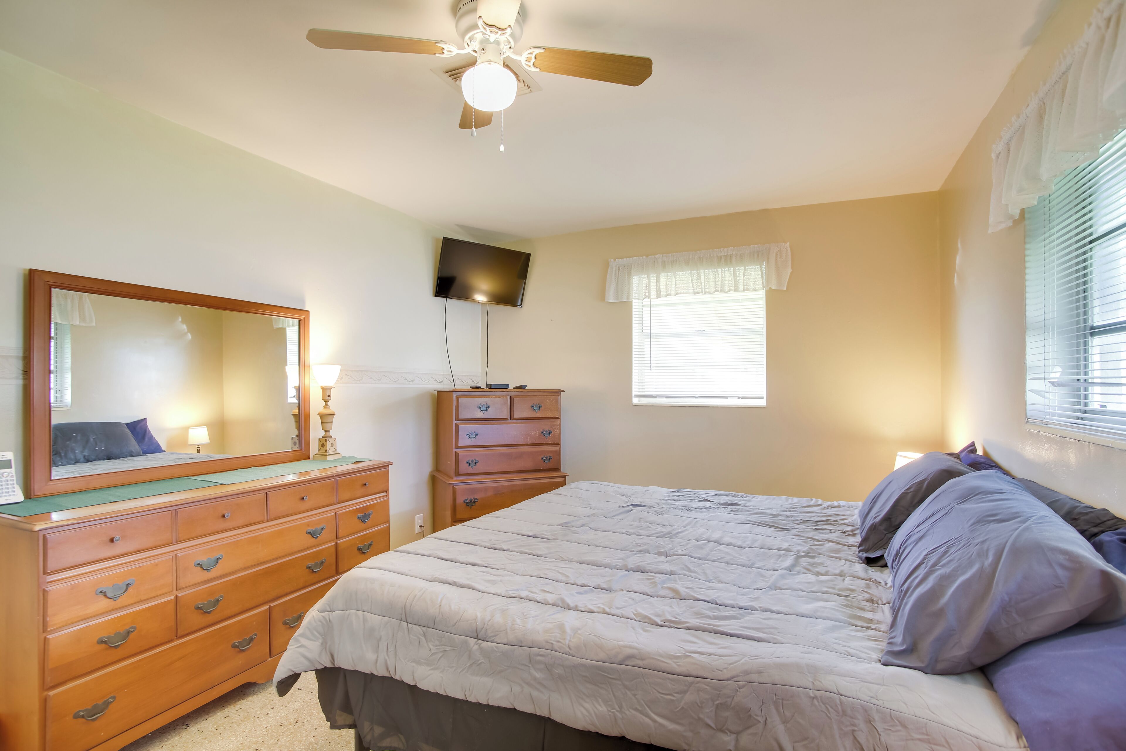 A bedroom with a bed, dresser, mirror, ceiling fan, and window with blinds.