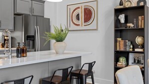 Una cocina moderna con armarios grises, un refrigerador de acero inoxidable, una encimera blanca y una planta en maceta. Hay dos taburetes de bar y una silla blanca.