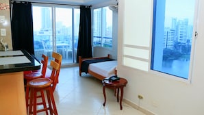 Ein Hotelzimmer mit einem Bett, einem kleinen Tisch mit Telefon, einem Sessel, einem Schreibtisch mit Stuhl und einem großen Fenster mit Blick auf die Stadt.