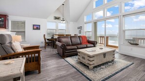 A spacious living room with a high ceiling, large windows offering ocean views, a brown leather sofa, a wooden coffee table, and a dining area with a wooden table and chairs.