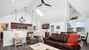 A spacious living area with a brown leather sofa, a wooden coffee table, and a dining table with chairs. A kitchen with modern appliances and wooden cabinets is visible in the background.