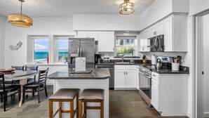 A modern kitchen with white cabinets, a central island with stools, a refrigerator, and a view of the ocean through a window. - Boundless Ocean Views From Home and Hot Tub, Pet Friendly, Private Deck, Fireplace, Steps to Beach (Depoe Bay)