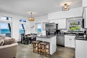 A modern kitchen with white cabinets, a stainless steel refrigerator, and a dining area with a view of the ocean.