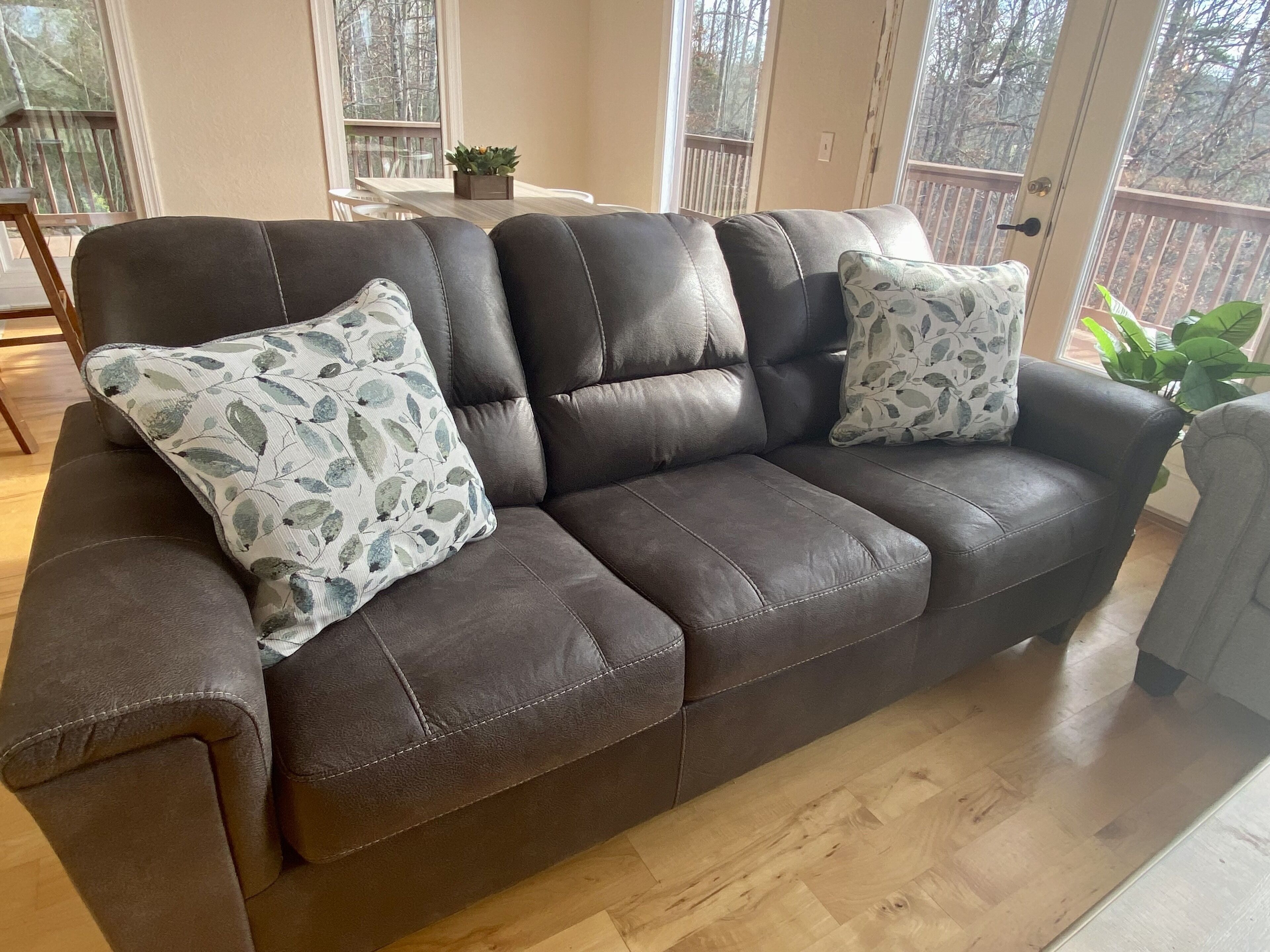 A brown leather sofa with floral cushions, positioned in a room with wooden flooring and large windows.