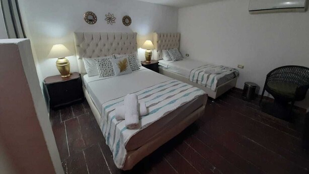 Quarto de hotel com duas camas, uma mesa de cabeceira com abajur, uma cadeira e um cesto de lixo pequeno.