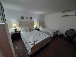 Habitación de hotel con dos camas, una mesita de noche con lámpara, una silla y un cesto de basura pequeño.