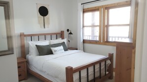 A bedroom with a wooden bed, white bedding, a bedside lamp, a wall-mounted circular decoration, and a window with blinds.
