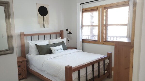 Un dormitorio con cama de madera, ropa de cama blanca, lámpara de noche, un adorno circular en la pared y una ventana con persianas.