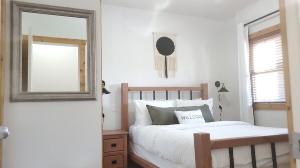 Ein Schlafzimmer mit einem hölzernen Bett, weißer Bettwäsche, einer Nachttischlampe, einem an der Wand befestigten Spiegel und einem Wandbehang aus Makramee.