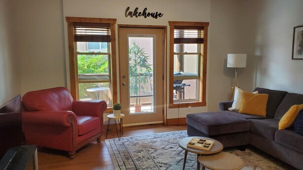 A living room with a red leather chair, a gray sofa with yellow pillows, a small round table, and a rug. There are three windows with blinds, and a sign above reads "Lakehouse".