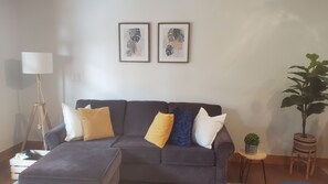Ein Wohnzimmer mit einem dunkelgrauen Sofa, mehreren Kissen, einer Stehlampe, zwei gerahmten Kunstwerken, einem kleinen runden Tisch mit einer Pflanze und einem Blumentopf neben dem Sofa.