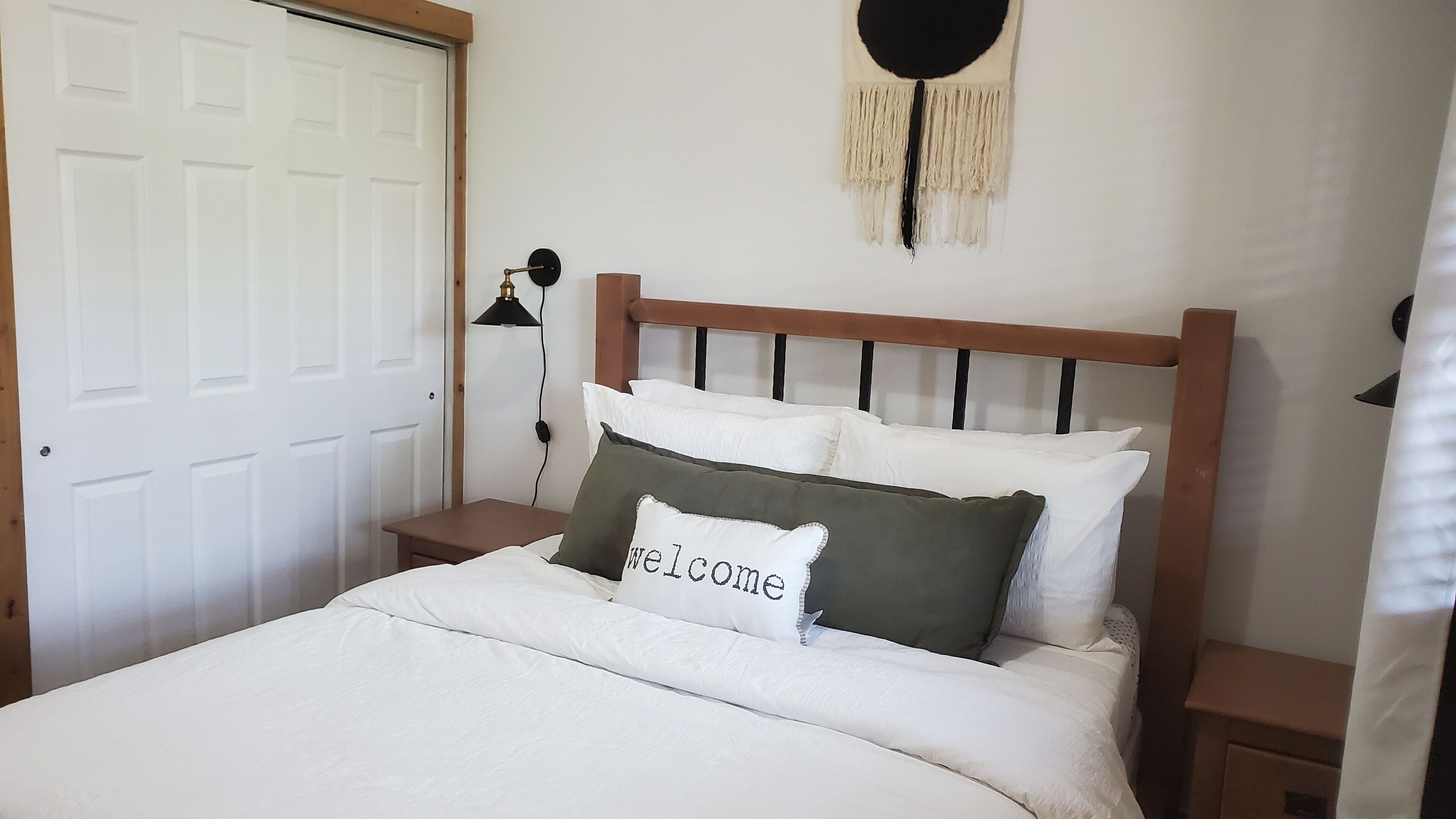 A neatly made bed with white bedding and a 'welcome' pillow, a wooden headboard, a bedside lamp, and a white door.