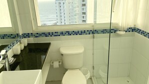 Une salle de bain moderne dotée d’une baignoire, de deux toilettes, d’une douche en verre et offrant une vue sur l’océan et les bâtiments.