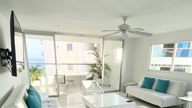 Un salon moderne avec un canapé blanc, une table basse et un fauteuil blanc. Il y a un balcon avec vue sur la mer.