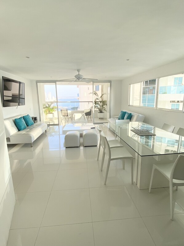 A modern living room with a white sofa, ottomans, a glass coffee table, and a TV mounted on the wall.