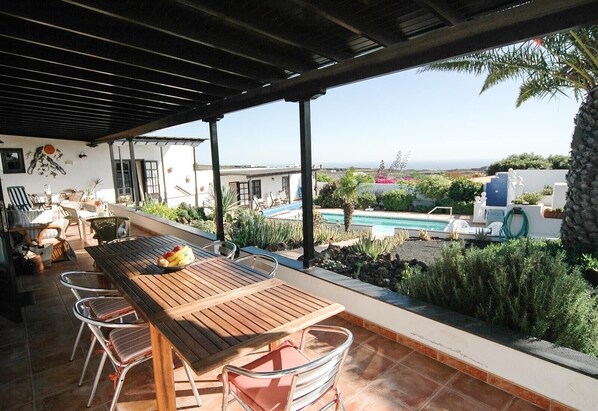 A covered patio with a wooden table and chairs, a pool, and a view of the landscape.