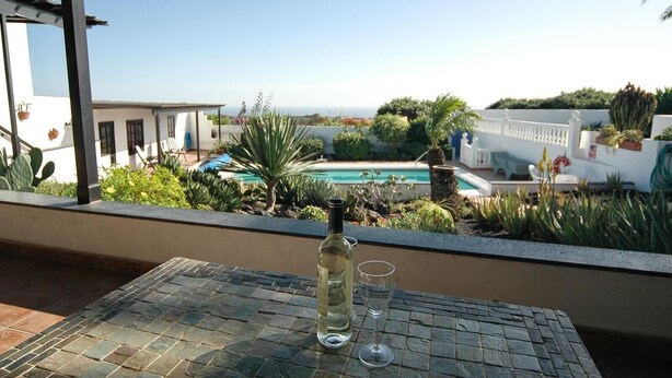 Una terraza con una mesa, una botella y una copa, con vistas a una piscina y un jardín.