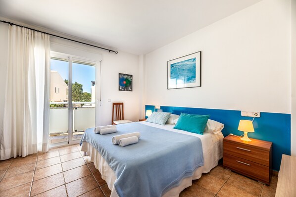 Habitación de hotel con cama, mesita de noche, lámpara, ventana con cortinas, un cuadro en la pared y vistas a edificios.