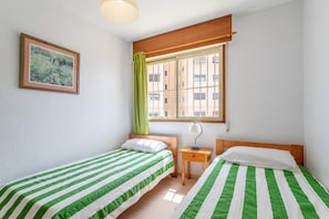 A bedroom with two beds, a window with a view of buildings, a painting on the wall, and a small bedside table with a lamp.