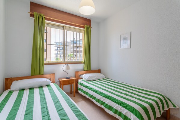 Une chambre avec deux lits simples, une petite table, une lampe, une fenêtre avec des rideaux et un tableau encadré au mur.