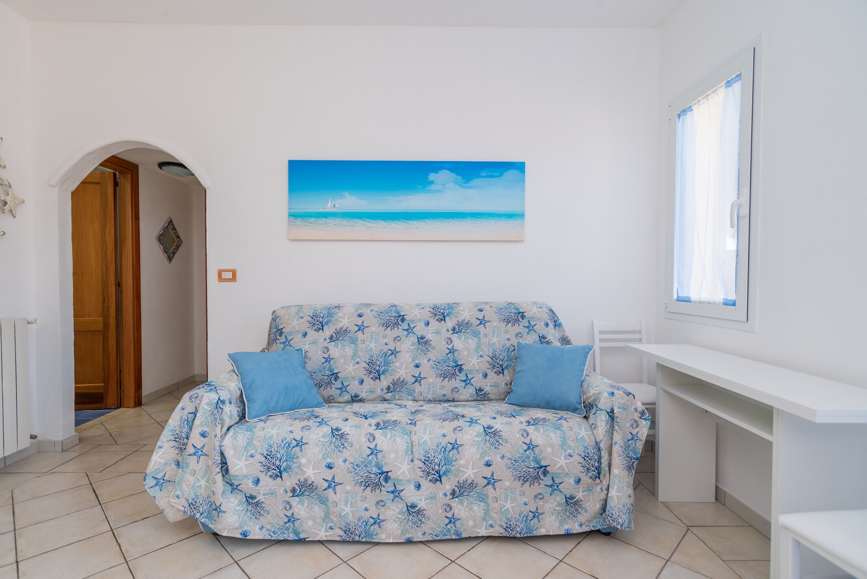 Una stanza con un divano decorato, un quadro raffigurante una spiaggia, una porta in legno e una finestra con tende.