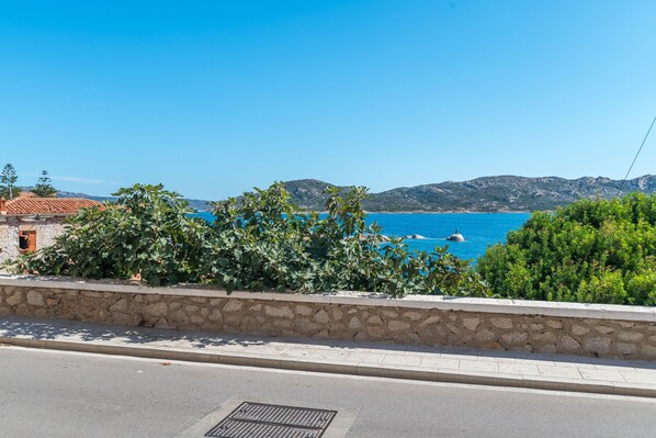 Une route côtière bordée d’un mur de pierres, une mer d’un bleu limpide et des collines à l’horizon.