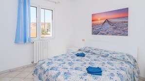 Ein Schlafzimmer mit Bett, Fenster mit Vorhängen, Heizkörper und einem Sonnenuntergangsbild an der Wand.
