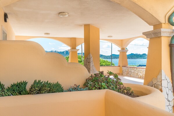 A covered outdoor area with a view of the sea, featuring a curved wall, columns, and plants.