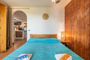 Une chambre à coucher avec une tête de lit en bois, un lit recouvert d’une couverture à rayures bleues, une table de chevet avec une lampe, et, au fond, une vue sur une cuisine.