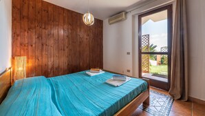 Een slaapkamer met een houten wand, een bed met een blauwe spreideken, een hanglamp en een deur naar een buitenruimte.