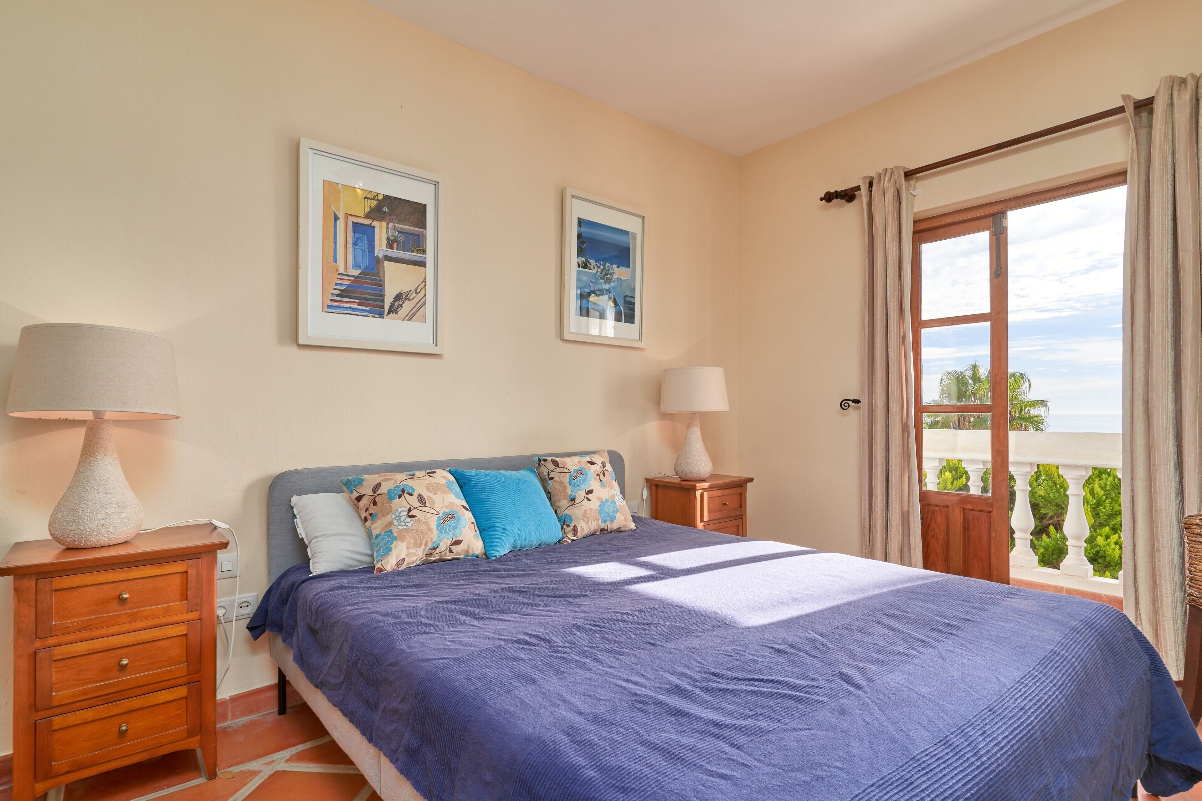 Un dormitorio con cama, dos mesitas de noche, un ventanal con cortinas y dos cuadros en la pared.