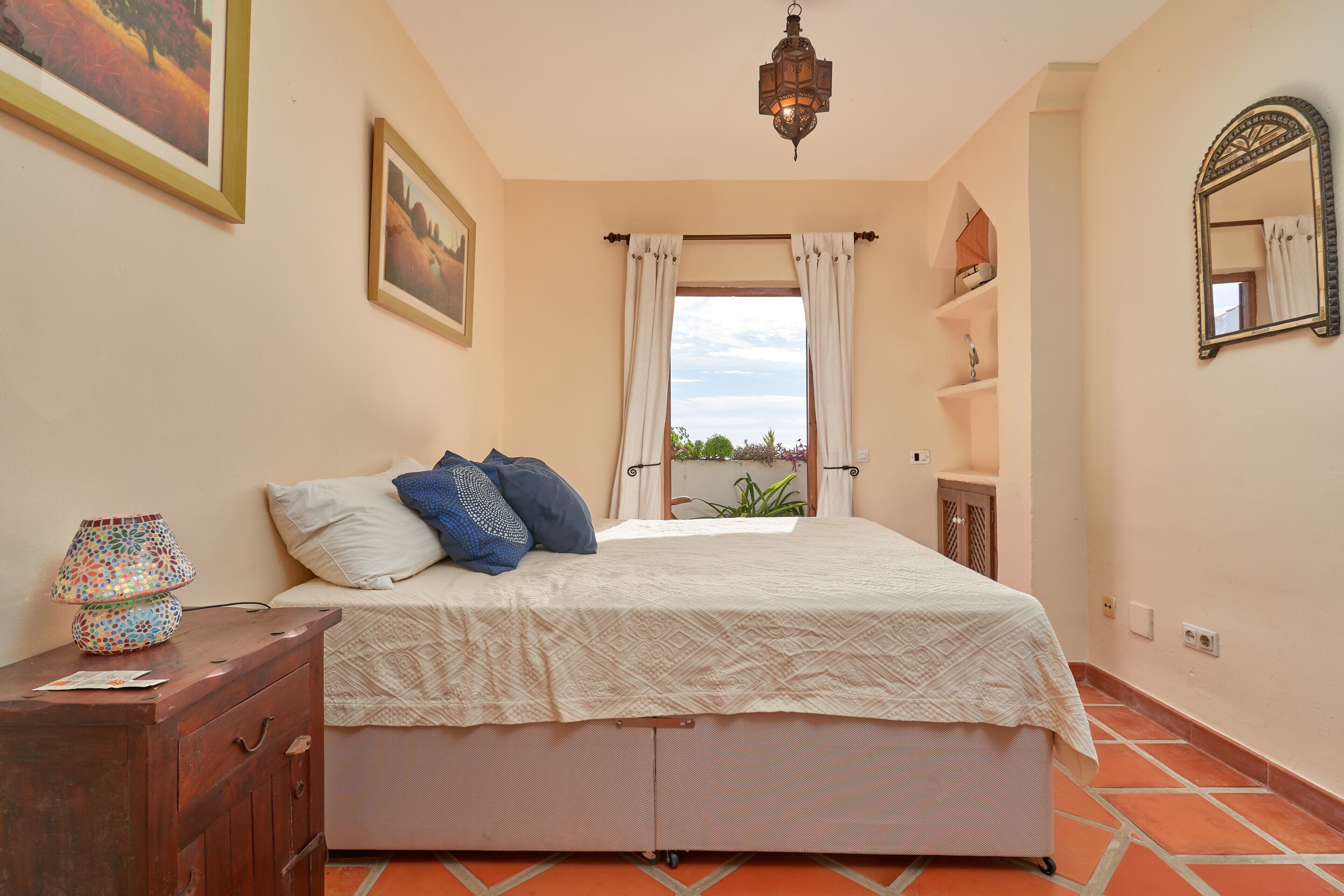 A bedroom with a bed, a dresser, a lamp, a window with curtains, a shelf, and a mirror.