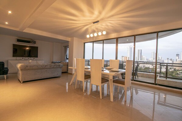 A modern living room with a sofa, a dining table with chairs, a TV mounted on the wall, and large windows offering a city view.