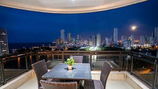 A balcony with a table and chairs, overlooking a city skyline at night. - Luxury 8 Bedroom Penthouse with Private Jacuzzi (Cartagena)