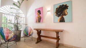 Een kamer met een houten bank, twee kleurrijke stoelen, een potplant en twee ingelijste kunstwerken aan de muur.