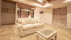 Una sala de estar con un sofá color beige, una mesa de centro, vigas de madera en el techo, una ventana con contraventanas y una pared de piedra.