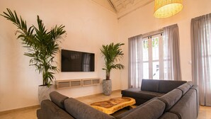 A modern living room with a sectional sofa, a wooden coffee table, two large potted plants, a flat-screen TV, and a large window with curtains.