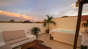 A rooftop terrace with a white sofa, a wooden coffee table, a hot tub, and a view of the city skyline.