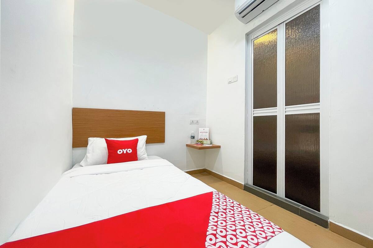 A hotel room with a bed, a wooden headboard, a red and white bedspread, a small bedside table with a bottle and a box, and a sliding glass door with a view.