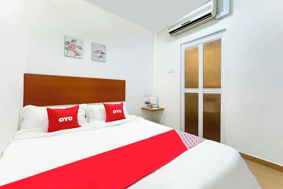 A hotel room with a large bed, a wooden headboard, two red pillows with "OYO" branding, a white bedspread, and a red throw blanket. There is a small table with a bottle and a glass, and a window with a view of the outside.