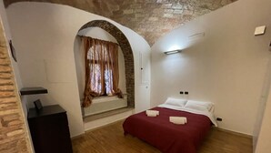 A bedroom with a bed, a desk, a window with curtains, and a brick wall.
