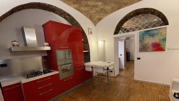 A compact kitchen with red cabinetry, a gas stove, and a microwave. There is a small dining area with a white table and chairs. The room has a rustic brick ceiling and white walls.