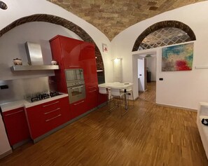 A compact kitchen with red cabinetry, a gas stove, and a microwave. There is a small dining area with a white table and chairs. The room has a rustic brick ceiling and white walls. - San Lorenzo Guesthouse (Roma)